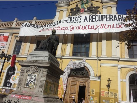Photo shows a building on the campus of the Universidad de Chile with a banner strung across it demanding free education. There is also a poster affixed to a pillar with a picture of Marx