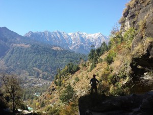 Mikey stands facing away from the camera. A valley falls down to a town. Beyond the town are green trees. Beyond the trees are snow-capped mountains.