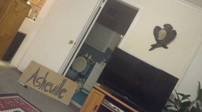 A large cardboard sign leans up against a wall dividing a bathroom and a living room that reads "Asheville" in permanent marker.