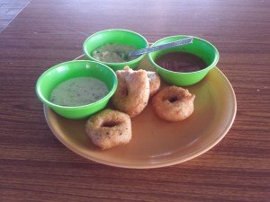 A plate has 3 small bowls on it filled with different colored indian gravies. There's also 4 fried vada on the plate.