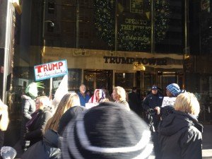 Several people stand around an enterance to a building with a sign that reads "TRUMP TOWER." One person holds a sign that reads "TRUMP. Make America Hate Again." A uniformed gangster stands nearby with a hat that reads "NYPD."