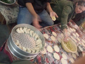 A table has many circular dumpling sheets scattered on it. In the middle is a bowl of filling. Near the camera is a tray of momos ready for steaming.