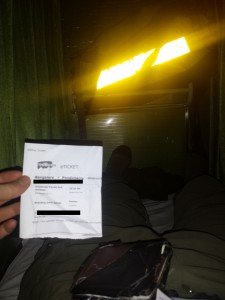 Mikey is laying in a bed taking a point-of-view shot. HIs wallet is on his chest, and his feet are stretched out in a bed. He holds a paper that reads "redBus.in eTicket, Bangalore -> Pondicherry"