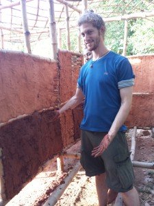 Mikey is smiling for the camera. He is wearing shorts, and his hands are covered in brown mud. In front of him is a wall made of panels of mud. Other panels can be seen covered in mud--some are exposed, showing a lattice of bamboo beneath the mud. A girl is standing just behind Michael. Above them is a bamboo lattice roof.