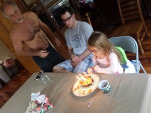 A young girl sits at a table in front of a plate of pancakes with lit candles in them. A man sits next to her looking on, and another man stands by them.