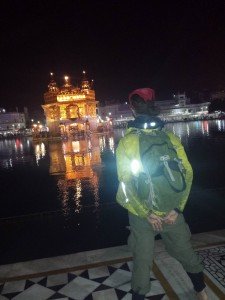 Mikey faces away from the camera. He is barefoot and his head is covered with a red bandanna. In front of him is a pool of water. In the middle of the water is a majestic Golden Temple.