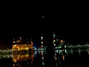 A body of water is shown at night with reflections of the scene above. In the middle of the water is a golden temple. Behind it there are two towers, and above the towers the full moon is in the night sky.