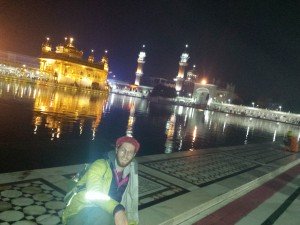 Mikey sits facing the camera, smiling. His head is covered with a red bandanna. Behind him is a pool of water. In the middle of the water is a majestic Golden Temple.