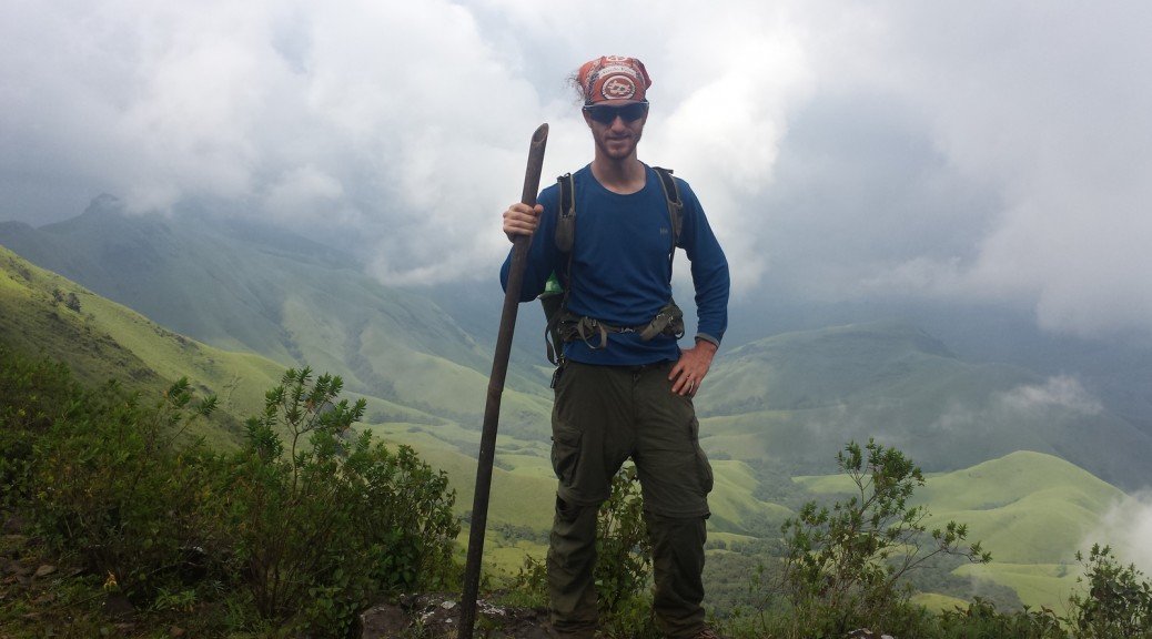 Mikey is seen holding a bamboo trekking pole and wearing a daypack with an orange bitcoin bandanna. In the background are lush green, rolling hills.