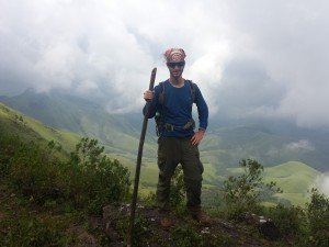 Mikey is seen holding a bamboo trekking pole and wearing a daypack with an orange bitcoin bandanna. In the background are lush green, rolling hills.