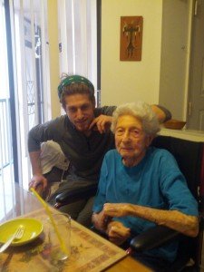 Mikey smiles. In front of him is an old woman sitting in a wheelchair at a dining table in her home.