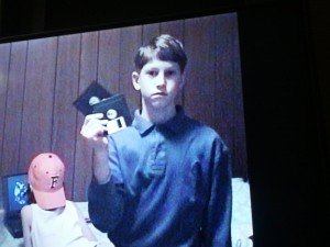 Mikey as a child wearing a buttoned-up collared shirt and holding up 2 floppy disks. Behind him, a girl sits at an old laptop wearing a hat labeled "F" for Florida.