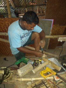An Indian man is seen holding a wood chisel in his hand. He is cutting into a short length of bamboo, which is propped on a table and held in-place with his toes. The table also has various carpentry instruments on it, including a drill, wooden drill bits, a vice, and a hand saw. Behind the man is are bamboo in all shapes and forms (ie: long, cut short, and latticed).