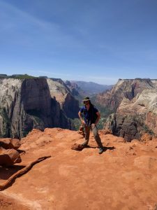 Mikey is smiling, standing near a cliff on top of a canyon. A valley between canyons stretches along in the distance behind him