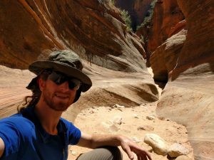 Mikey sits in a dry canyon bottom. The canyon's walls have been shaped by wind & rain into a beautiful winding shape.