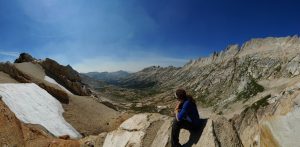 Mikey sits atop a mountain pass looking down into the valley below. The reddish-brown rock has patches of snow on the pass, and the valley below is green.