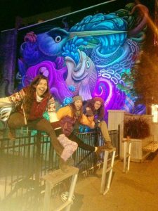 Mikey and two women are posing while standing on a fence. The wall behind them is a mural--very psychedelic and colorful--showing a bird, a fish, and an octopus.