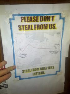 A sign hangs on the wall reading "PLEASE DON'T STEAL FROM US. STEAL FROM CHAPTERS INSTEAD." The sign includes a map to walk to the Chapters bookstore (to steal from).