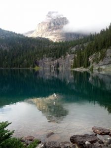 A mirror-like lake reflects a tall mountain behind it. Around the lake are lush green trees.