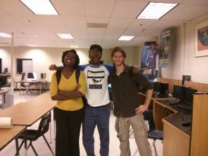 3 people embrase each other, smiling for the camera, in a classroom with many, many computers.