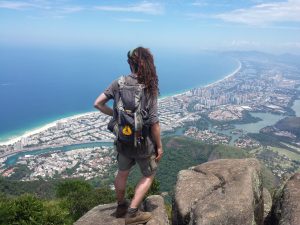 Mikey stands on a rocky cliff overlooking a jungle juxtiposed next to a city with a canal running through it, and emptying-out to the ocean, which stretches out to the horizon.