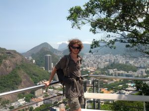 Mikey smiles facing the camera while leaning up against a railing. Behind him is a mix of jungle, skyscraper skyline, and tall rocky mountains.