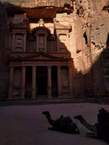 A huge, majestic facade carved out of a sandstone cliff is seen. In front of it, two camels lay in the sand.