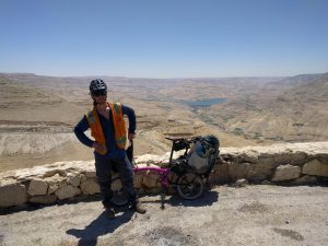 Mikey stands triumphantly with his bicycle leaned against a short rock wall. Behind him is Wadi Mujib. The landscape is brown desert with patches of irrigated green. In the distance, the Mujib Dam and its huge 35 million cubic meter water reservoir can be seen.