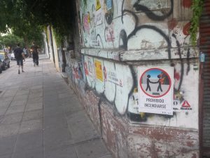 A street sidewalk adjorns a concrete wall full of graffiti, stickers, and signs. One sign has a picture reminiscent of the album cover for "Wish You Were Here" by Pink Floyd, and it has the caption "Prohibido Incendiarse."