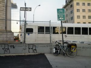 Mikey's bicycle leans up against a sign reading "Manhattan Bridge" with a picture of a bicycle on it. Behind a concrete barrier covered in graffiti, a white bus with dark tinted windows drives past with the inscription "Brooklyn Transportation Corp"