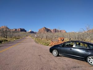 Mikey's car is parked in the shoulder in front of a sign that reads "Zion National Park Kolob Terrace." There are empty trees on either side of the road, and tall canyons in the distance.