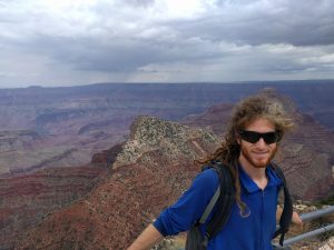 Mikey is smiling, standing against a railing. Behind him are reddish canyons that make up the Grand Canyon.