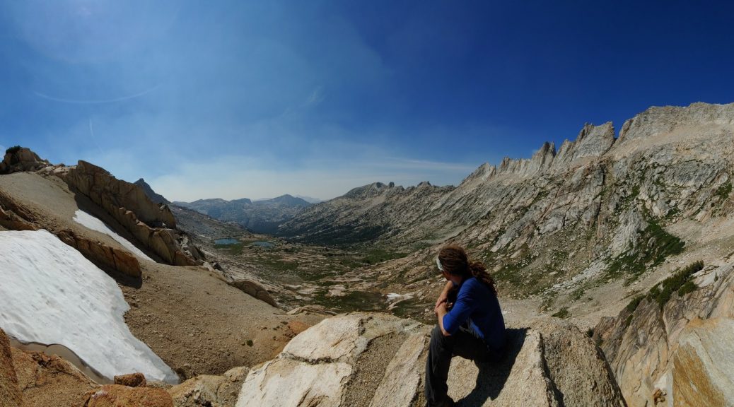 Mikey sits atop a mountain pass looking down into the valley below. The reddish-brown rock has patches of snow on the pass, and the valley below is green.