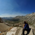 Mikey sits atop a mountain pass looking down into the valley below. The reddish-brown rock has patches of snow on the pass, and the valley below is green.