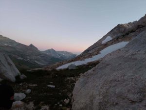 The sun is setting behind the rocky mountains in the high sierras. Near a snow patch in a meadow, a tent is setup.