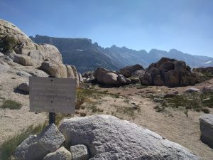 A sign propped-up by rocks in a mountain pass reads "ENTERING YOSEMITE WILDERNESS. WILDERNESS PERMIT REQUIRED. PETS AND FIREARMS PROHIBITED. FIRES AT DESIGNATED SITES ONLY. PACK OUT ALL THAT YOU PACK IN. BEAR HABITAT."
