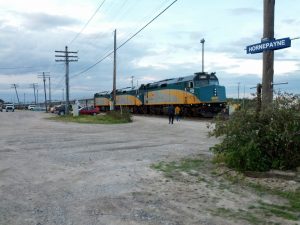 The engine of the Via Rail train is shown, and the passenger cars stretch back along the tracks as far as the eye can see. A small sign indicates that the stop is "Hornepayne, Canada"