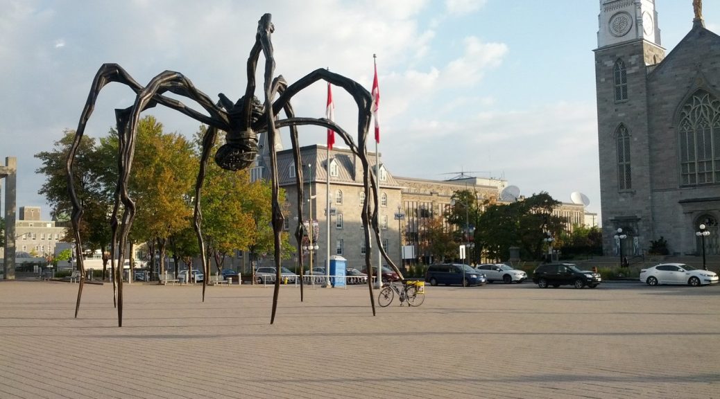 Mikey's bicycle is leaned-up against a towering, 30-ft tall sculpture of a spider (Maman) in Ottawa. Canadian flags are behind it. A church tower is seen to the right.