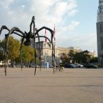 Mikey's bicycle is leaned-up against a towering, 30-ft tall sculpture of a spider (Maman) in Ottawa. Canadian flags are behind it. A church tower is seen to the right.