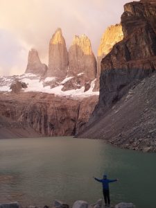 Mikey is seen facing the camera smiling with his arms raised in excitment. Behind him is a body of water and behind that are the Torres Del Paine towers. The tops of the towers are just beginning to be lit by the rising sun.