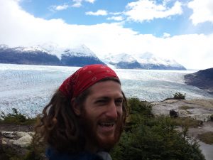 Mikey is smiling in a selfie. Behind him is a long valley. The entire valley is filled with an immense blue-white-grey glacier full of crevasses. Behind the glacier are snow-capped mountains.