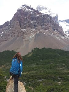 Mikey stands facing away from the camera. He is looking out at a lush green valley. The valley ends after a few kilometers as an enormous snow-capped mountain juts up from the ground.