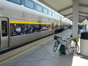 Mikey's bicycle is shown on a train platform with a train labeled "California Department of Transportation" and a logo on the train of a piece of luggage and a bicycle