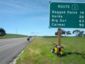 Mikey's bicycle rests against a sign indicating "Route 1" while J cycles past