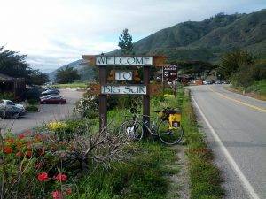 Mikey's bicycle is leaned up against a sign reading "Welcome to Big Sur" with lush, green hills in the background