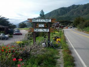 Mikey's bicycle is leaned up against a sign reading "Welcome to Big Sur" with lush, green hills in the background