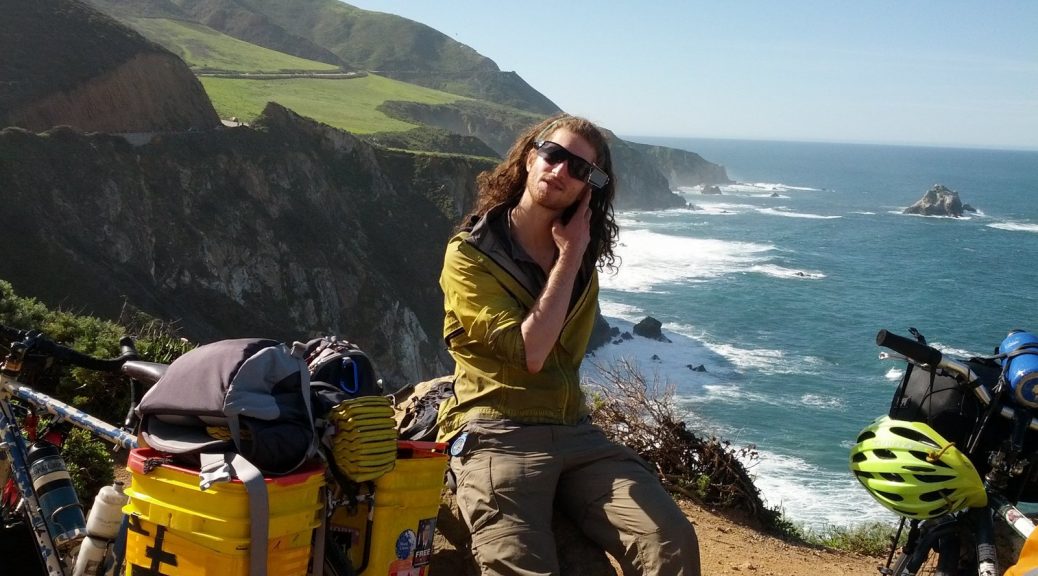 Mikey is shown sitting on a rock next to his bicycle. Behind him is a cliff overlooking the Pacific Ocean.