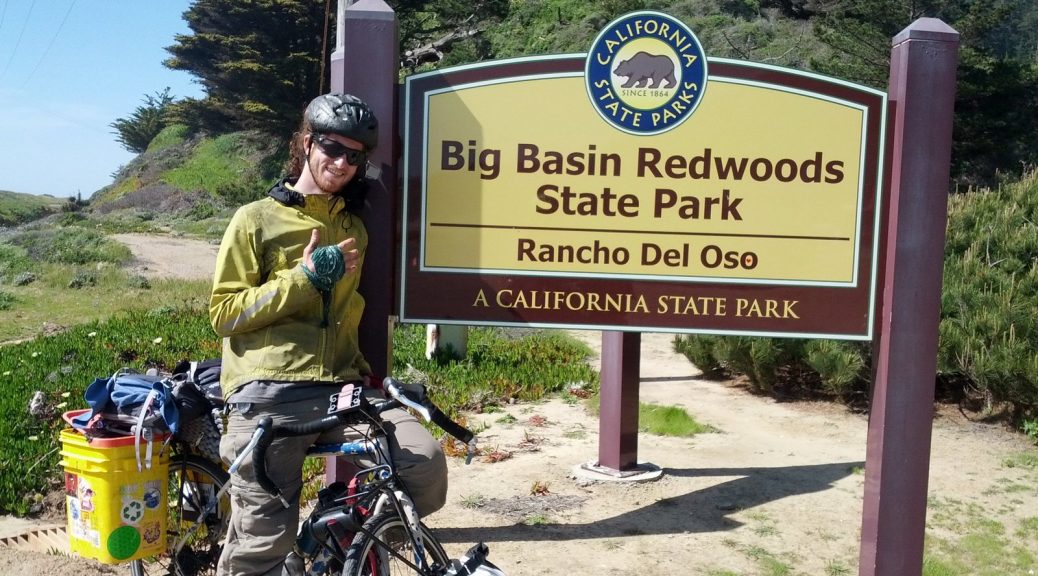 Mikey is smiling, pointing to a sign that reads "Big Basin Redwood State Park, Rancho Del Oso, A California State Park"