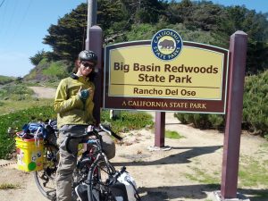 Mikey is smiling, pointing to a sign that reads "Big Basin Redwood State Park, Rancho Del Oso, A California State Park"