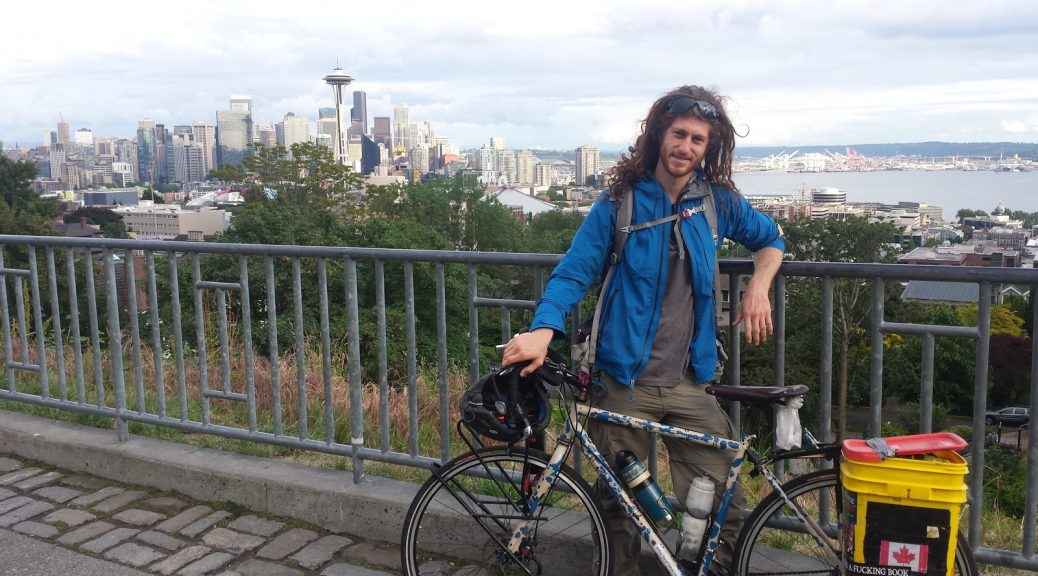 Mikey stands with his bicycle in front of a railing overlooking the Seattle Skyline and the Puget Sound